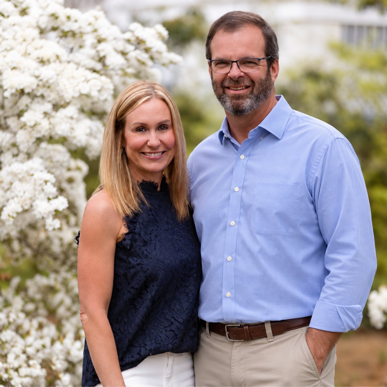Joe and Meaghan Perger standing side by side outdoors in front of a tree covered in white spring blossom, Meaghan in a dark sleeveless lace top and white trousers, Joe in a light blue button-down shirt, glasses and a grey beard