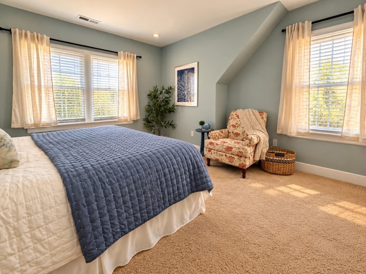 A sunlit bedroom with freshly painted pale blue-green walls, white trim and beige carpet, with a blue quilted bed, a floral armchair and two tall windows in cream curtains